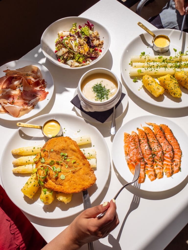 A table set with plates of smoked salmon, schnitzel with white asparagus and sauce, prosciutto, a grain salad, and a bowl of creamy soup, with hands holding forks and sunlight streaming in.