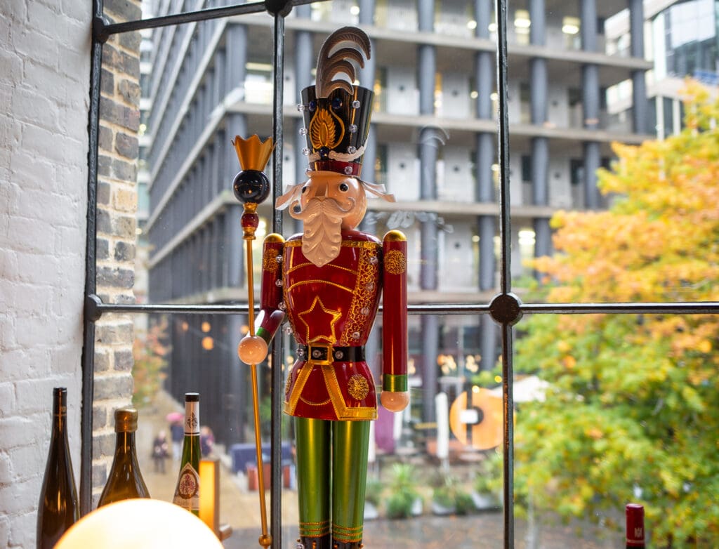 A colourful wooden nutcracker soldier stands on a windowsill beside wine bottles, with modern buildings and autumn trees visible through the large window behind it.