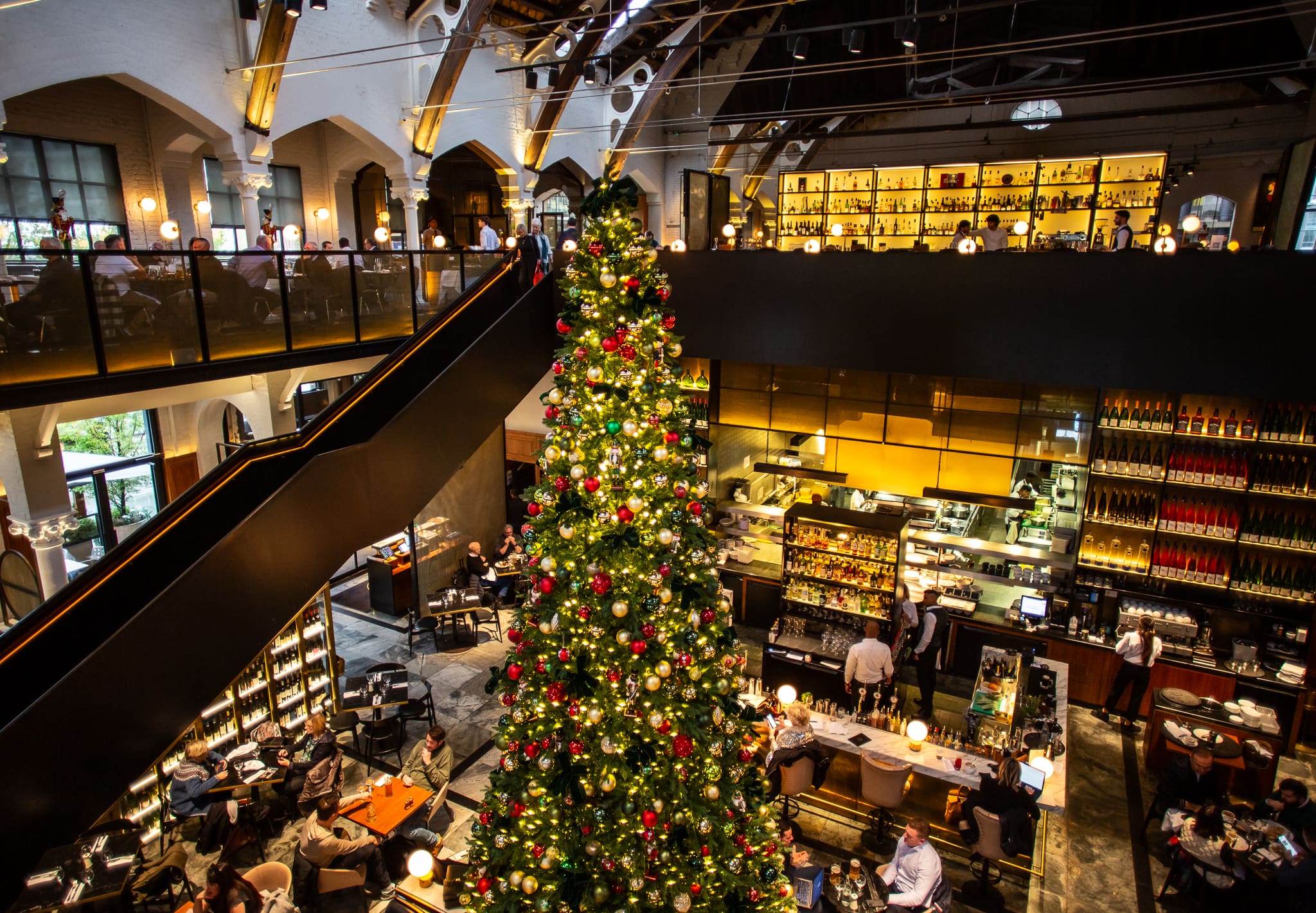 A large, decorated Christmas tree stands in the centre of a spacious, modern restaurant with high ceilings, balcony seating, and warm lighting. People are dining at tables on both levels.