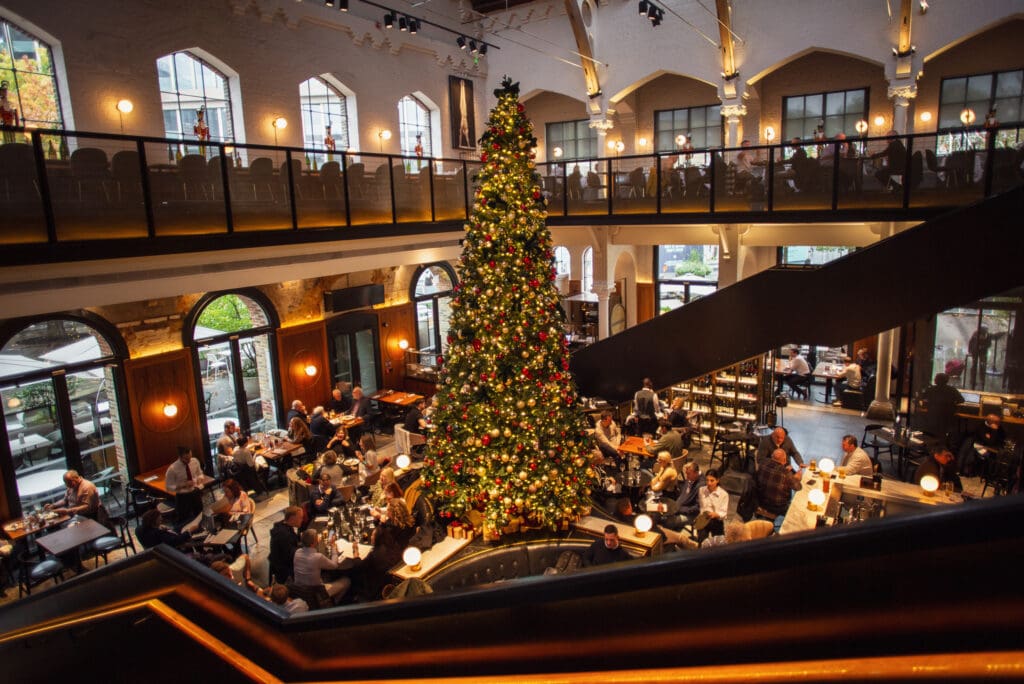 A large, decorated Christmas tree stands in the centre of a busy restaurant with high ceilings, large windows, and diners seated at tables on both the ground floor and mezzanine level. Warm lighting creates a festive atmosphere.