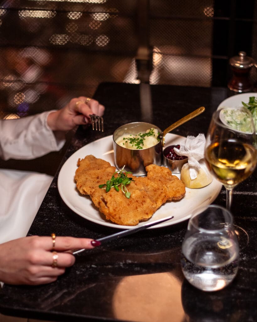 A person dining at a restaurant with a plate of breaded fried schnitzel, mashed potatoes with chives, a lemon wedge, cranberry sauce, and a glass of white wine on a dark table.