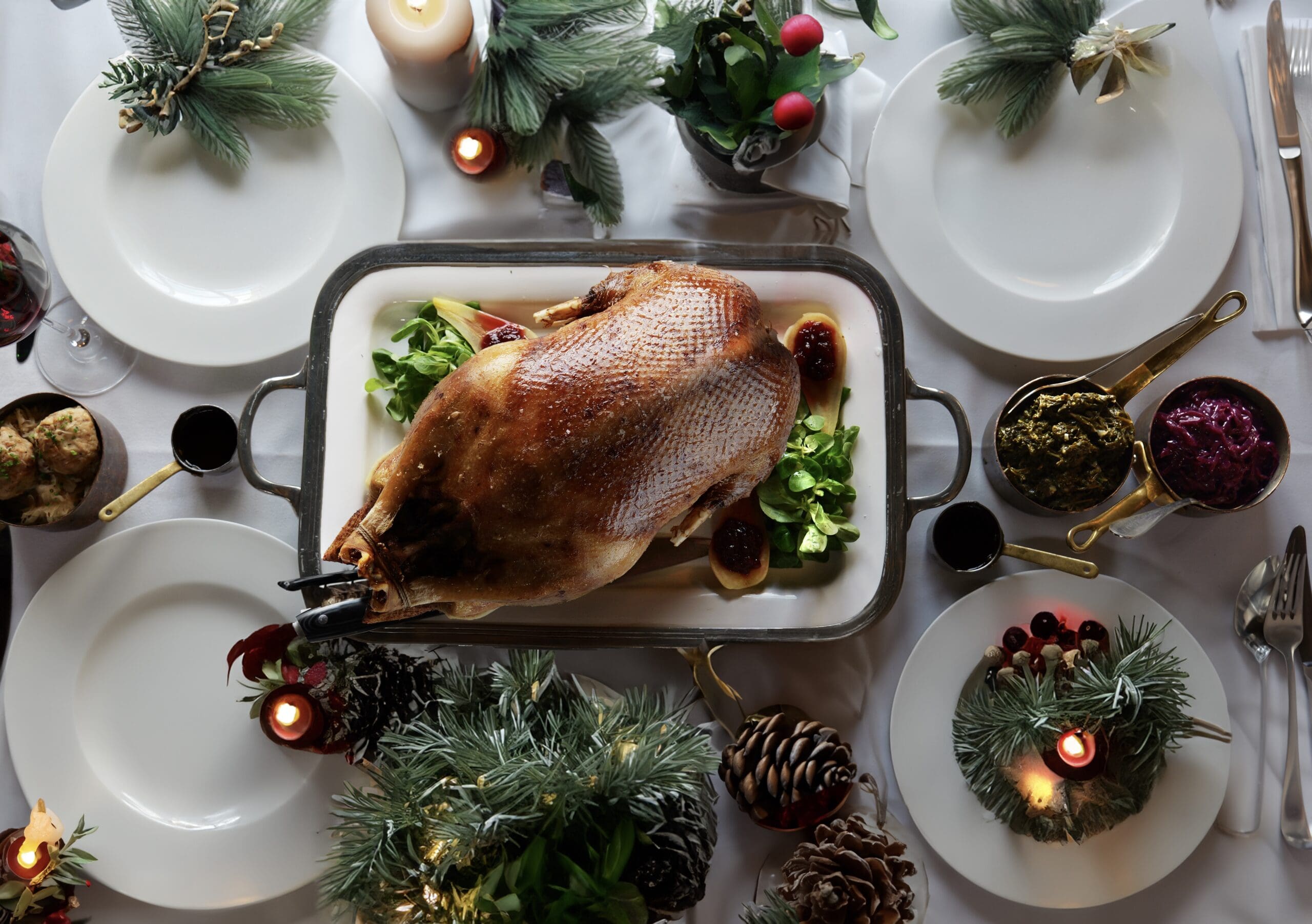 A roasted whole bird on a platter surrounded by greens sits at the centre of a festively decorated holiday table with candles, pine branches, pinecones, and plates set for four. Various side dishes are also present.