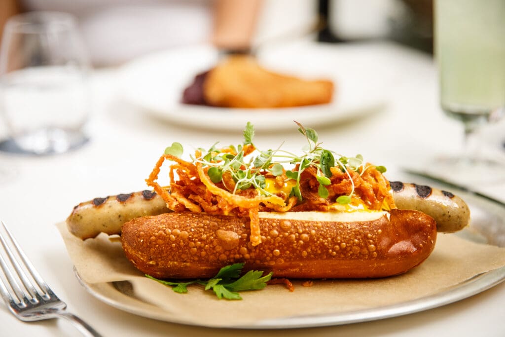 A grilled sausage in a pretzel bun topped with fried onions and microgreens is served on a piece of greaseproof paper, with a fork and knife nearby on a white tablecloth.