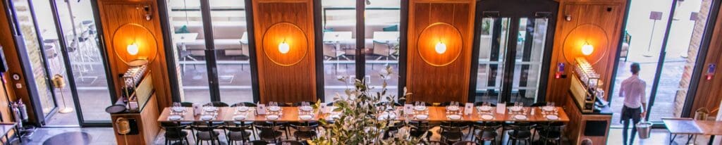 A modern restaurant interior with wooden walls, round lights, and a long dining table set with plates and glasses. Large windows and glass doors let in natural light. A person stands near the entrance.