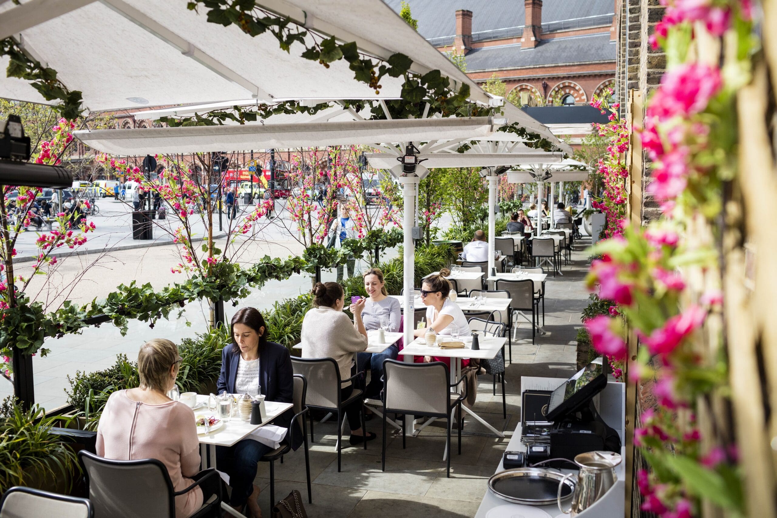 People sit at outdoor café tables under white umbrellas, surrounded by pink flowers and green vines, enjoying food and conversation on a sunny day. A busy street and brick buildings are visible in the background.