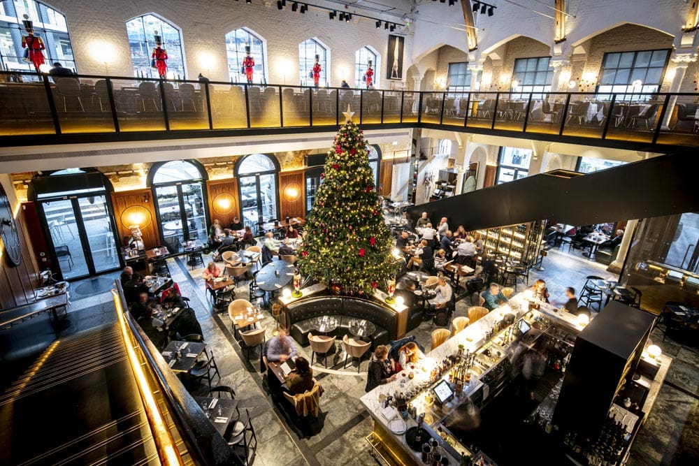 A large, elegant restaurant decorated for Christmas with a tall, decorated tree in the centre, festive lights, and many people dining at tables on both the ground floor and an upper balcony.