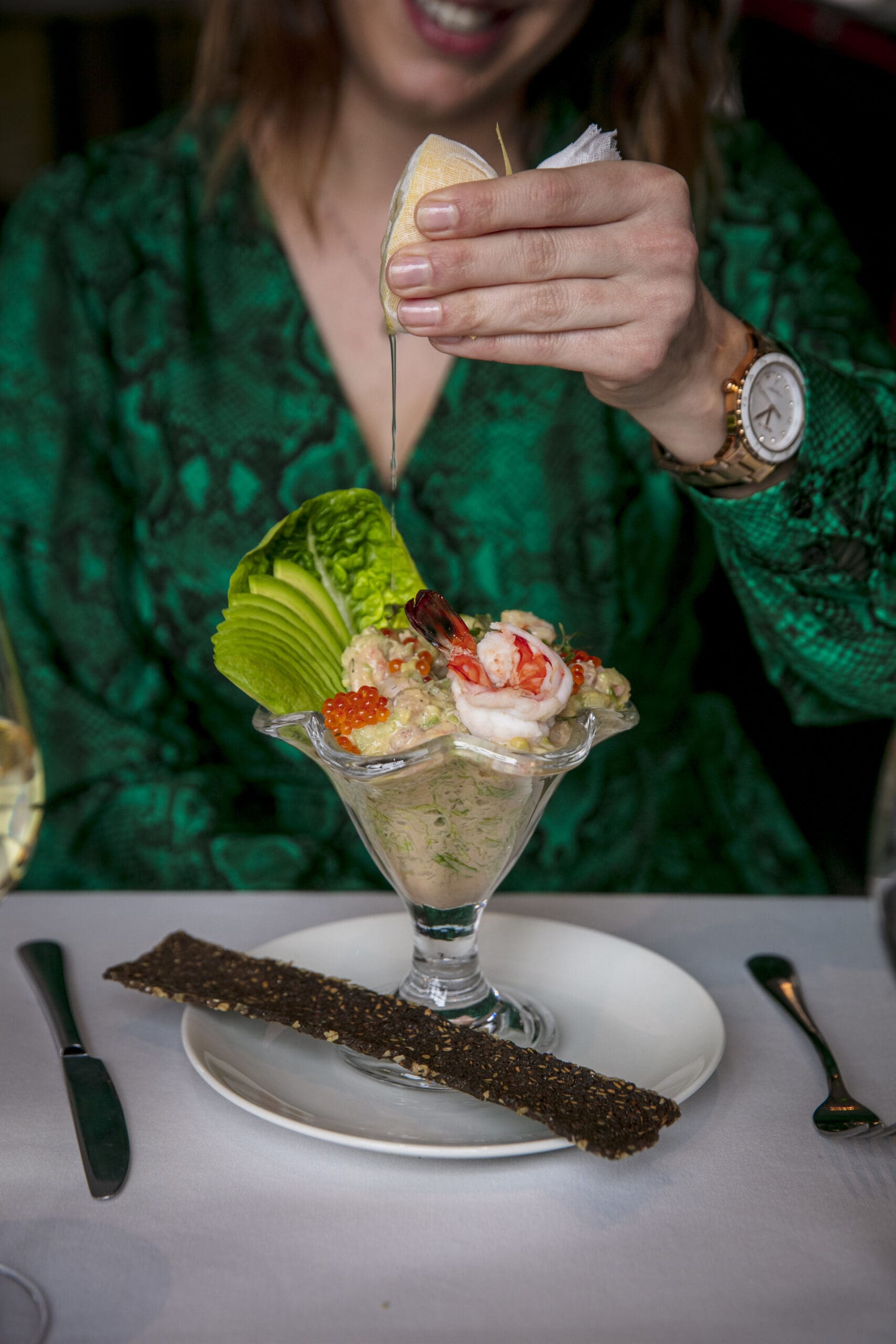 A person in a green outfit squeezes a lemon over a seafood cocktail topped with prawns, roe, and lettuce, served in a glass on a white plate with a crispbread on the side.