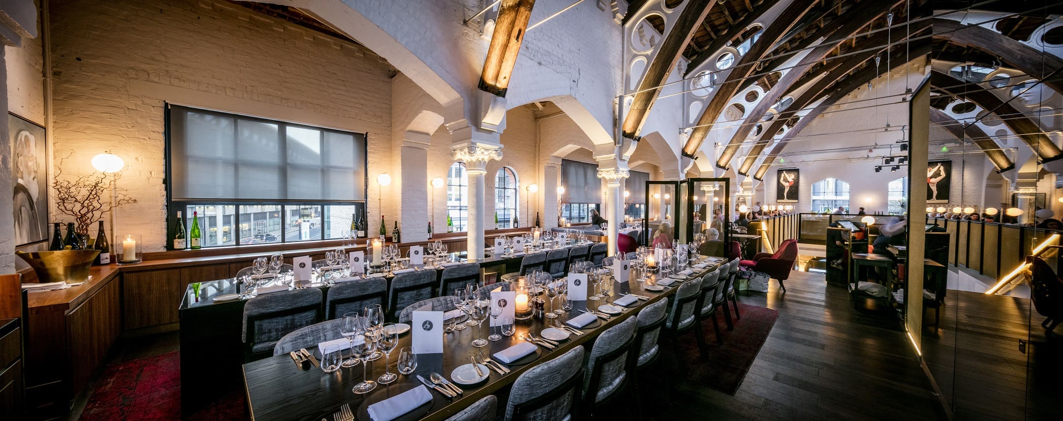 Elegant restaurant with long, set dining tables, stylish chairs, arched ceilings with exposed beams, and soft lighting. Large windows and tasteful decor create a sophisticated and inviting atmosphere.