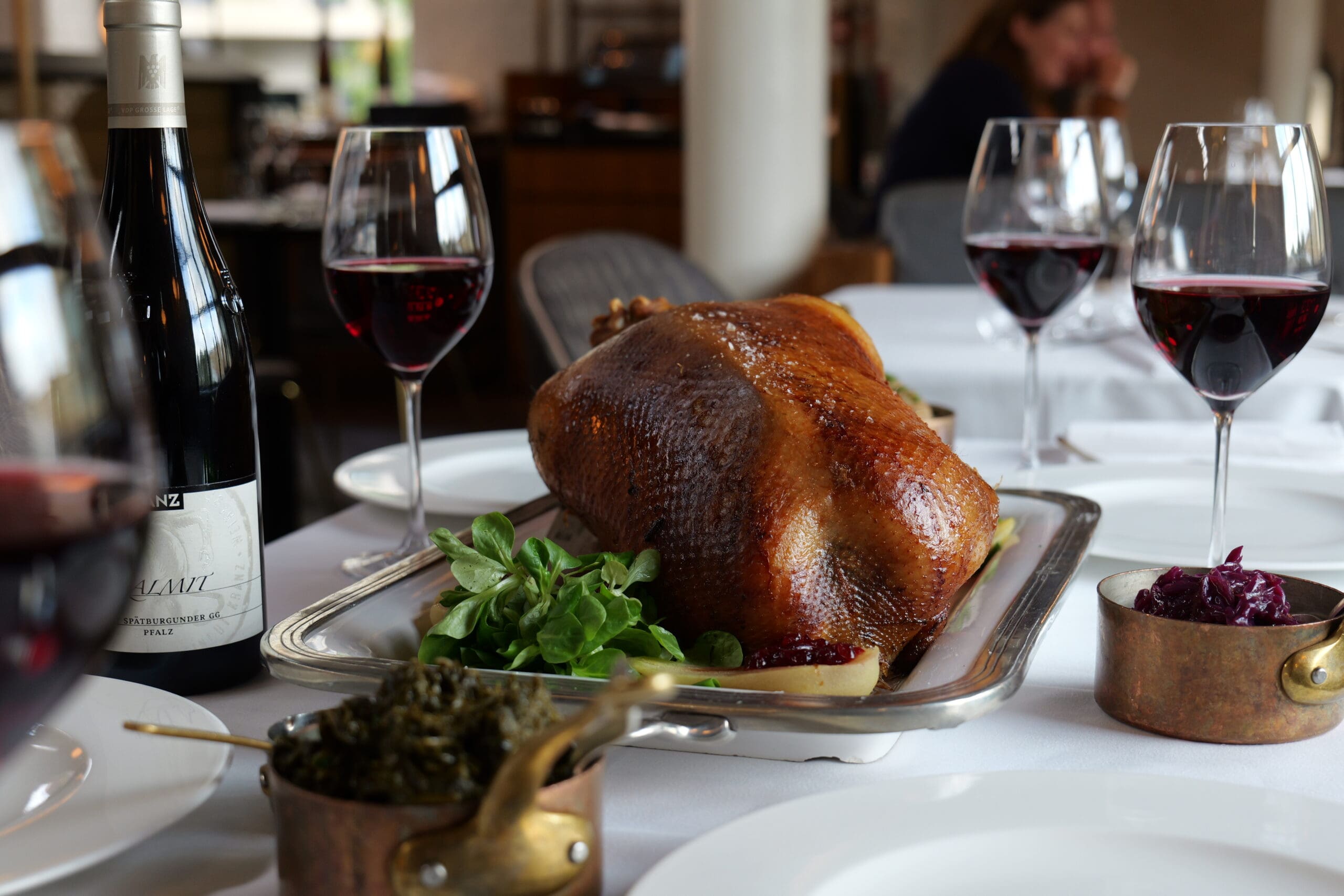 A roast goose on a platter is served with vegetables, surrounded by glasses of red wine, a wine bottle, and small pots of side dishes on a white tablecloth in an elegant restaurant setting.